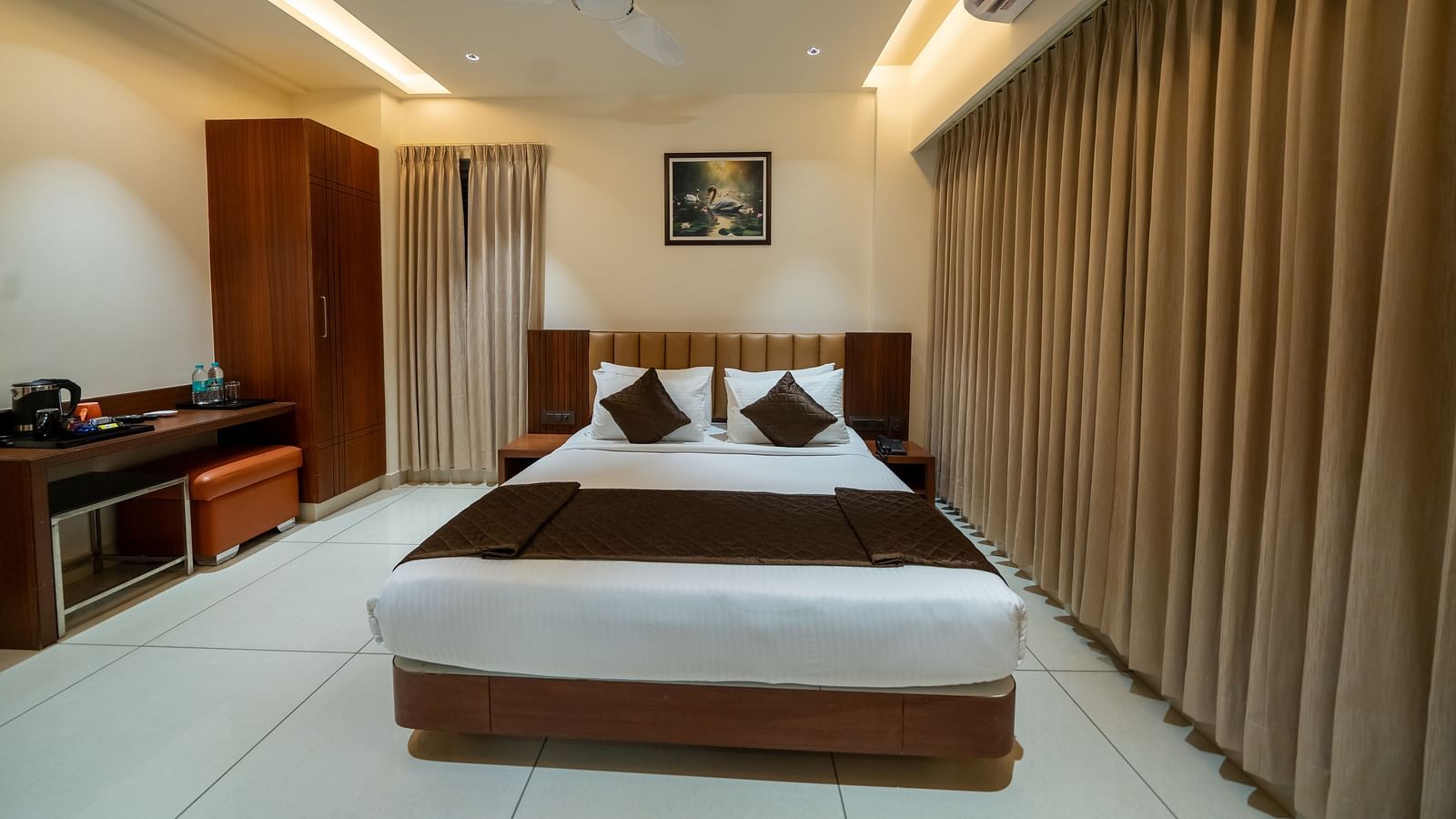 A front view of the bed at Hari Govinda's Express, Vijayanagar, with a photo frame above the headboard. To the left, a wooden cupboard stands beside a table with essentials, while on the right, an AC is mounted above the fully drawn curtains.