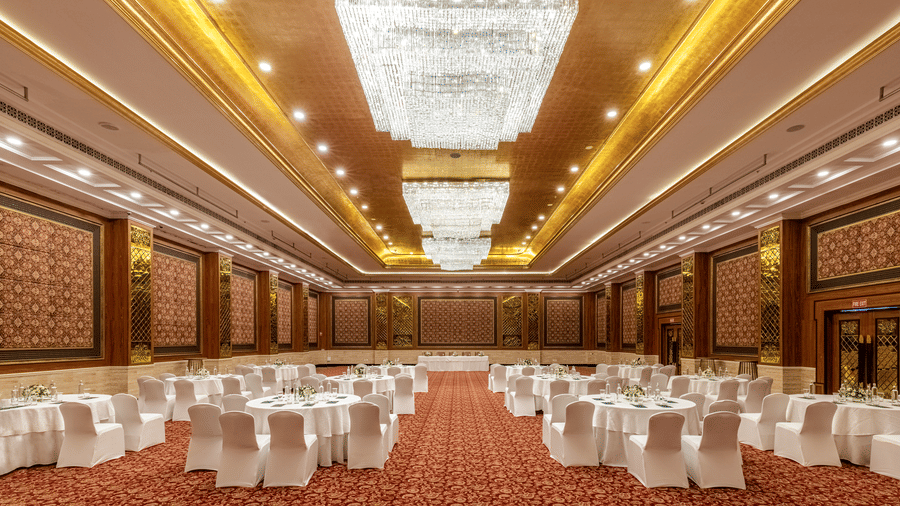 Ballroom featuring a large banquet hall at MAYFAIR Palm Beach Resort, Gopalpur-on-Sea. Chandeliers hang from the patterned ceiling. Rows of dining tables and chairs are arranged on the floor facing a stage area.