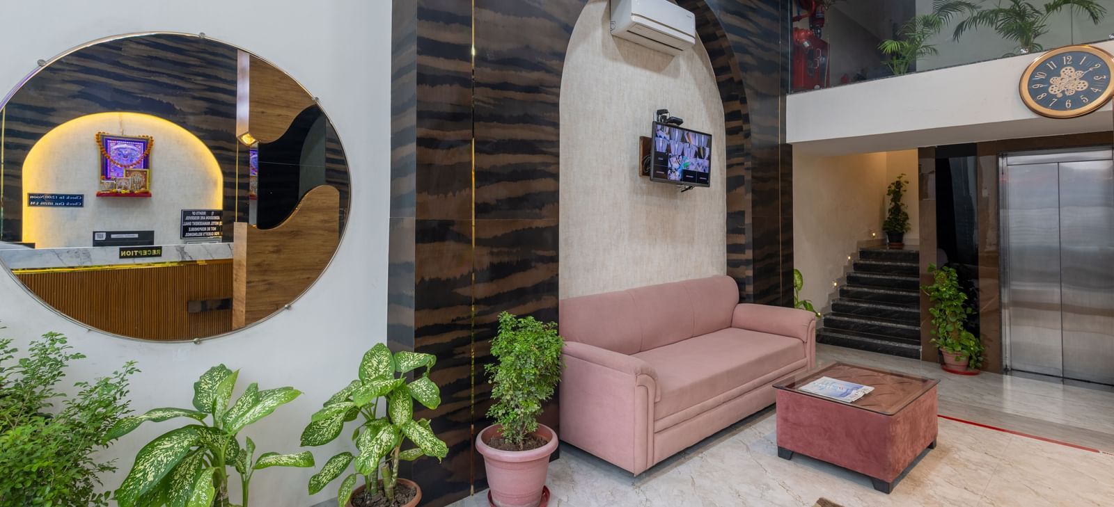 Lobby with round mirror, pink sofa, and potted plants at Pravasa Stay Dewas Naka, Indore.