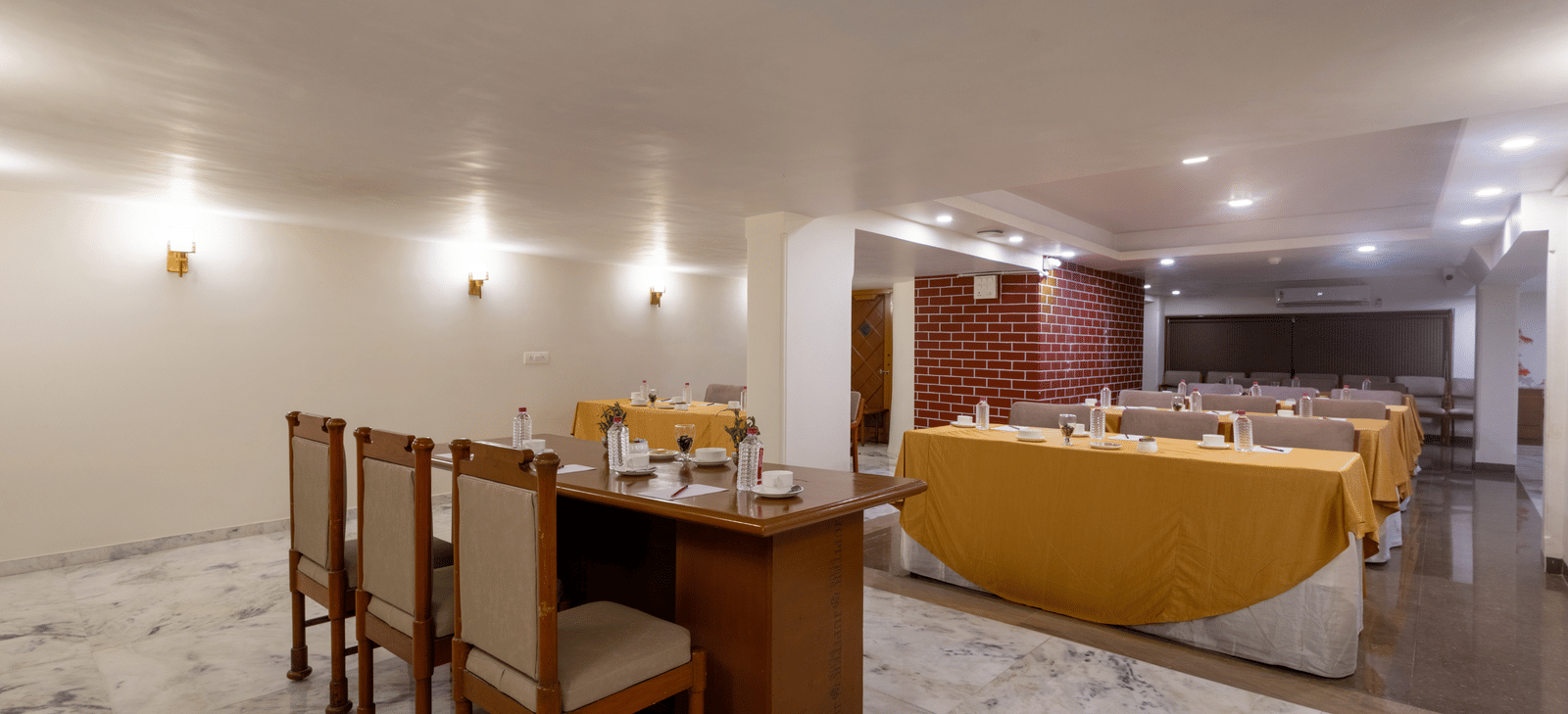 A well-lit hotel dining space showing a corner with wooden chairs, a table set for dining, and a yellow sofa at Pravasa Stay, Vadodara.