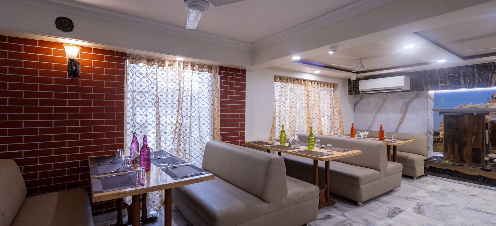 Corner of a restaurant with soft sofa seating, a coffee table, a ceiling fan, and red brick wall detailing at Pravasa Stay, Vadodara.