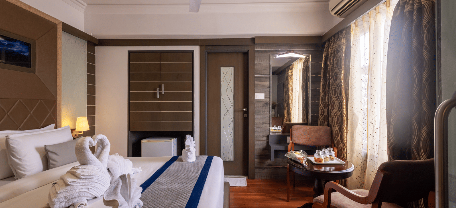 View of the bed reflected in a large mirror/television screen, highlighting the room's warm tones and wooden features at Pravasa Stay, Vadodara.