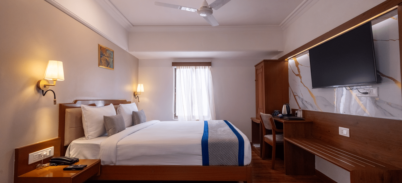 Close-up of the bed and a long wooden desk/console area with a television mounted above the mirror at Pravasa Stay, Vadodara.