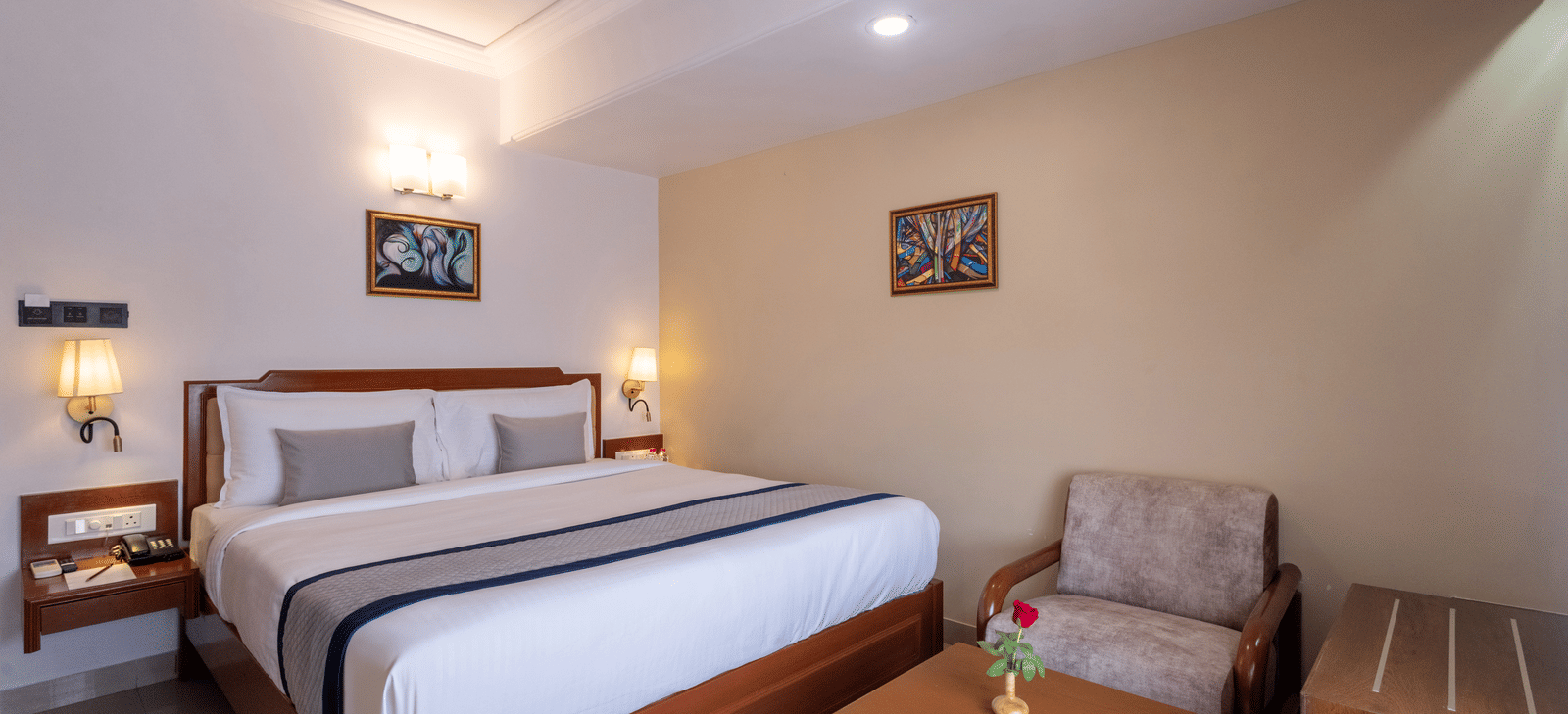 Close-up of a well-made hotel bed and a comfortable armchair with a side table, under ambient wall lights at Pravasa Stay, Vadodara.