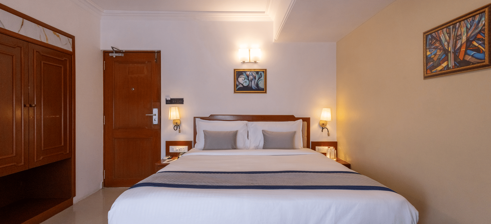 A view of the king-sized bed and headboard from the doorway, showing the wooden floor and warm bedroom lighting at Pravasa Stay, Vadodara.