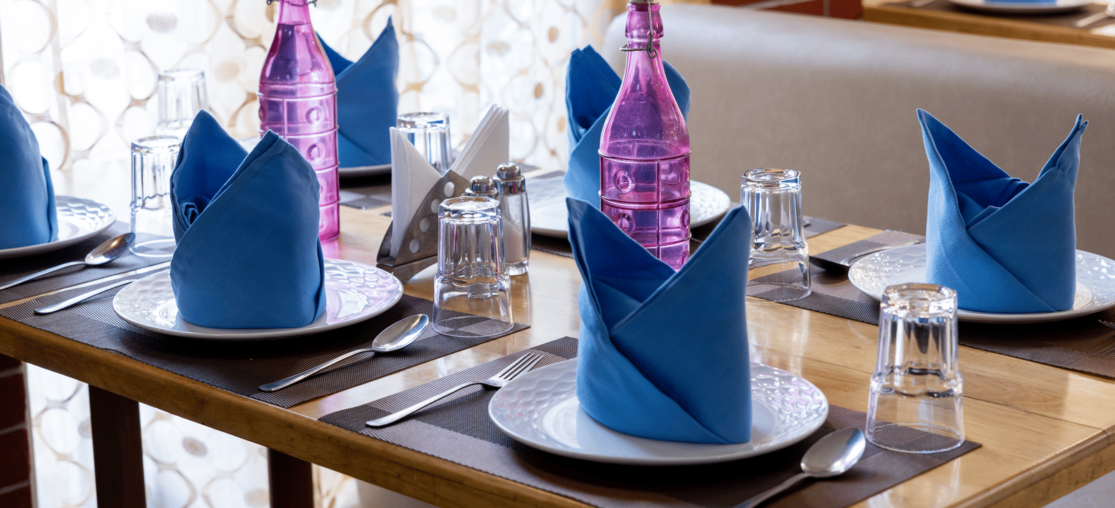 Close-up of a wooden table setting with white plates and striking blue and pink folded napkins in a well-lit dining area at Pravasa Stay, Vadodara.