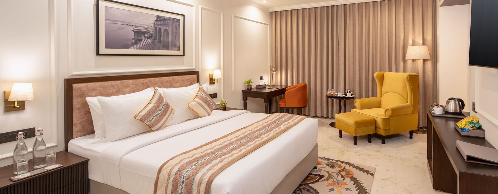 An image of a bedroom with bedside table, photo frame hanged on the wall, work desk with sofa chair, and a flat TV screen - Brij Eternity, Vrindavan