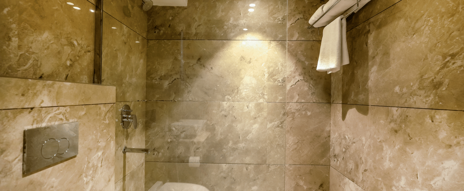 A modern, compact wet room or shower area clad in large, textured brown and beige marble-effect tiles at BluSalzz Select City Centre, Amritsar.
