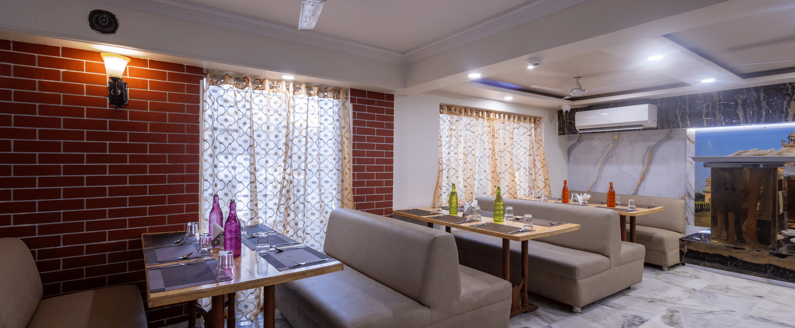 Corner of a restaurant with soft sofa seating, a coffee table, a ceiling fan, and red brick wall detailing at Pravasa Stay, Vadodara.