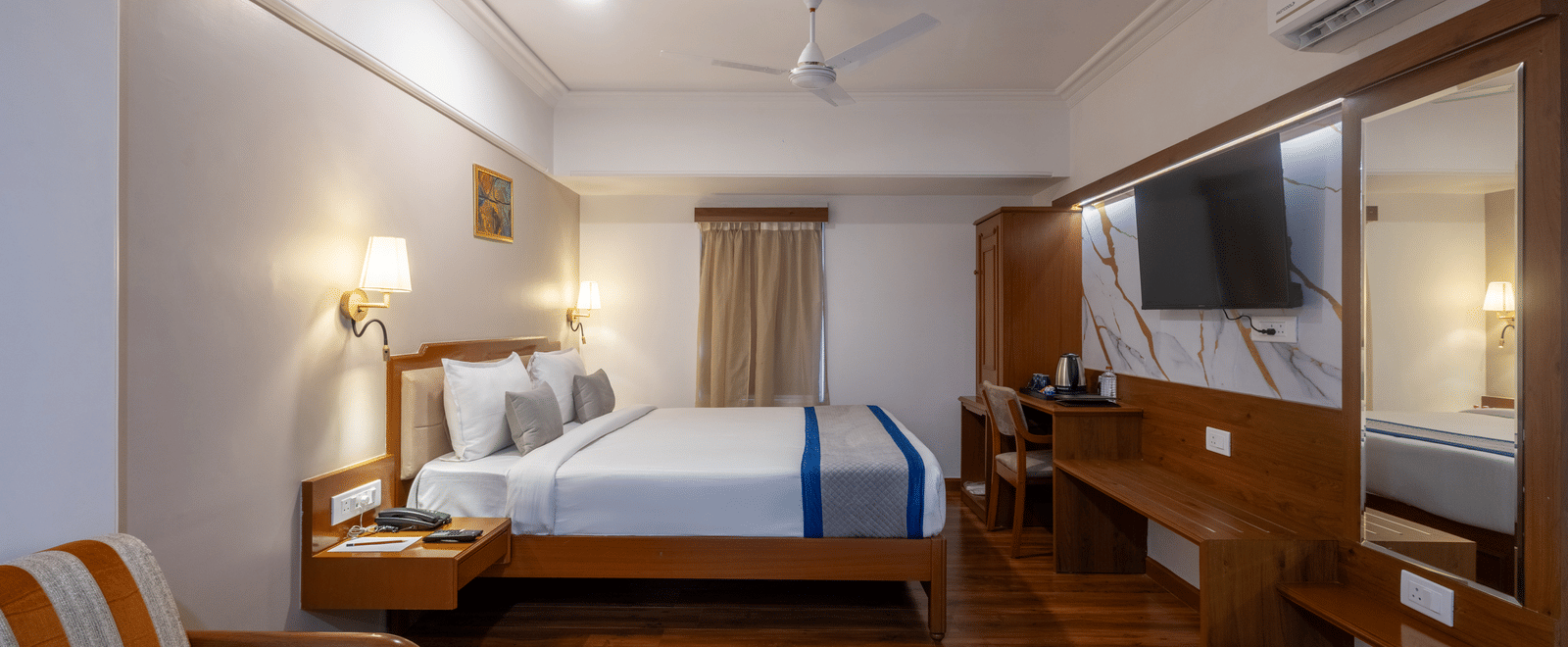 Wide view of a king-sized room showing the bed, built-in wooden furniture, and a full-length mirror behind the TV at Pravasa Stay, Vadodara.
