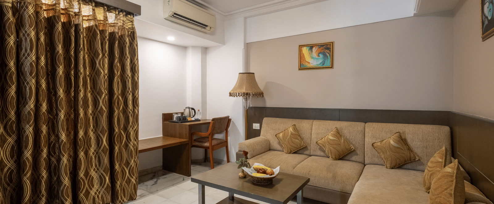 A hotel suite lounge with a large, beige L-shaped sofa, coffee table, and heavy, patterned brown curtains at Pravasa Stay, Vadodara.