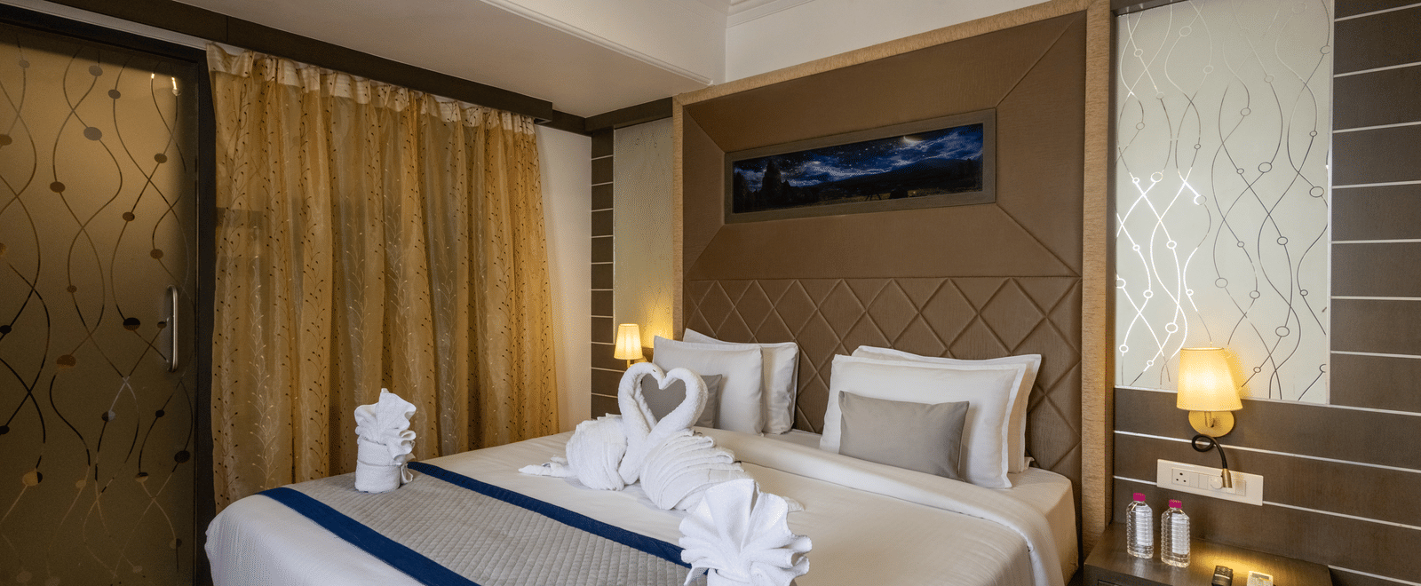 A hotel bedroom with a neatly made bed featuring towel swans and blue accents, wood panelling, and soft bedside lighting at Pravasa Stay, Vadodara.