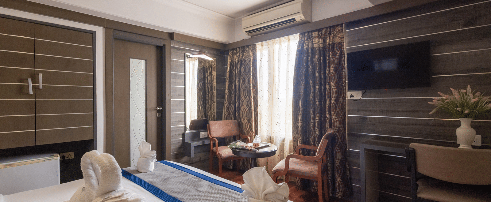 Wide view of a well-appointed hotel room showing the bed and a separate seating area near dark, patterned curtains at Pravasa Stay, Vadodara.