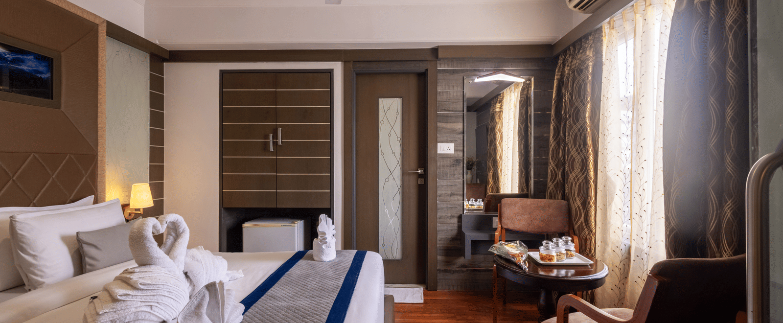 View of the bed reflected in a large mirror/television screen, highlighting the room's warm tones and wooden features at Pravasa Stay, Vadodara.