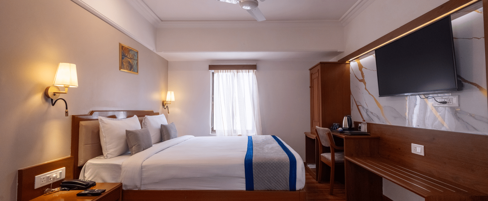 Close-up of the bed and a long wooden desk/console area with a television mounted above the mirror at Pravasa Stay, Vadodara.