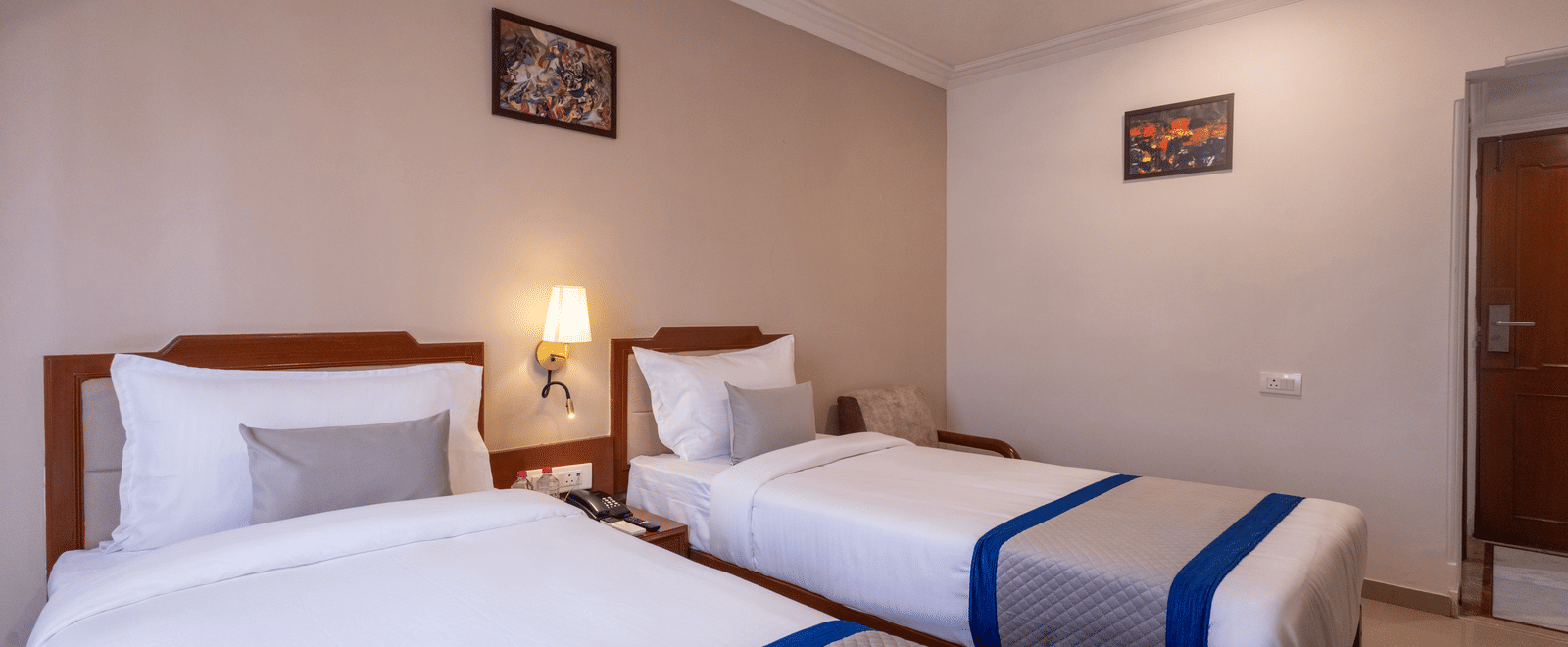 A bright hotel room with two single beds, each made up with white linen and a striking blue and grey throw at Pravasa Stay, Vadodara.