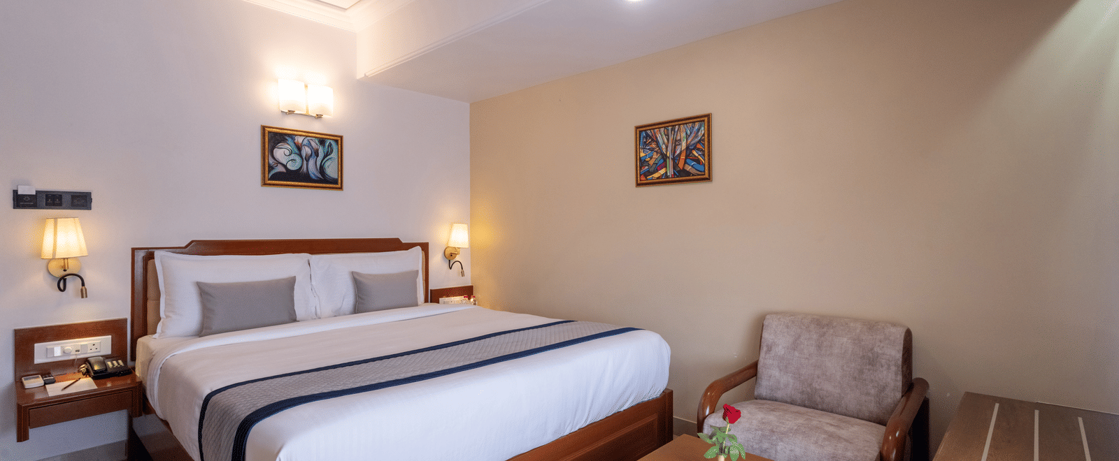 Close-up of a well-made hotel bed and a comfortable armchair with a side table, under ambient wall lights at Pravasa Stay, Vadodara.