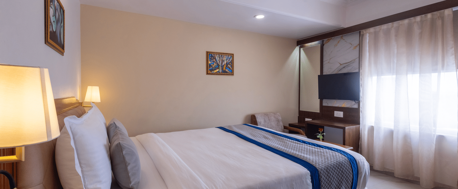 A warm, contemporary hotel room with a large bed and bright window, featuring wood accents and soft lighting at Pravasa Stay, Vadodara.