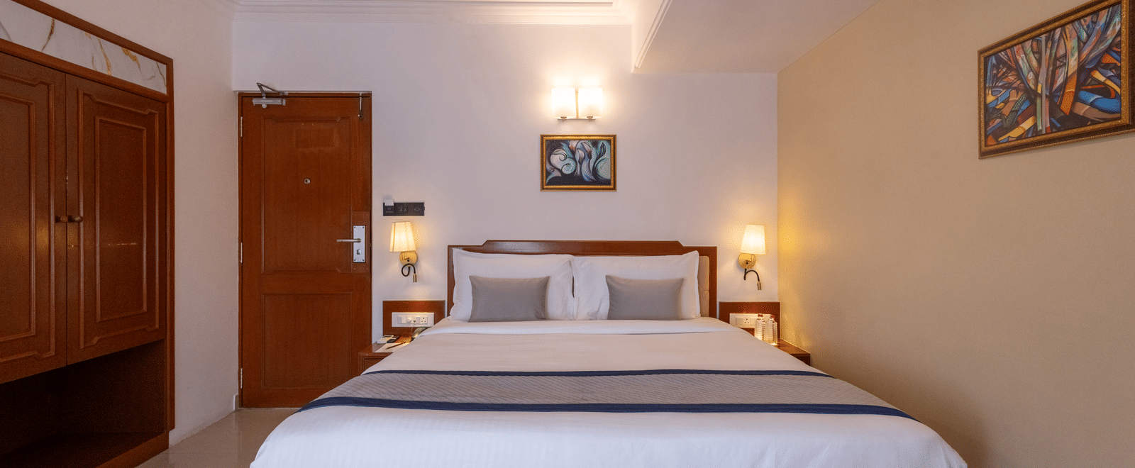 A view of the king-sized bed and headboard from the doorway, showing the wooden floor and warm bedroom lighting at Pravasa Stay, Vadodara.