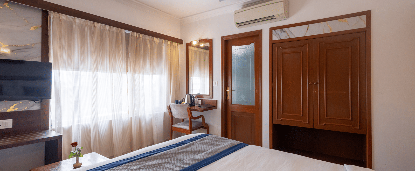 View across the bed towards a large wooden wardrobe and mirrored door, showing the room's spacious layout at Pravasa Stay, Vadodara.