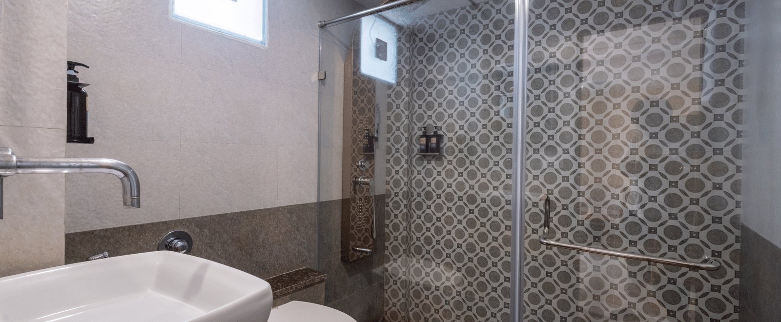 A sleek hotel bathroom shower area with a glass partition and decorative patterned grey and white wall tiles at Pravasa Stay, Vadodara.