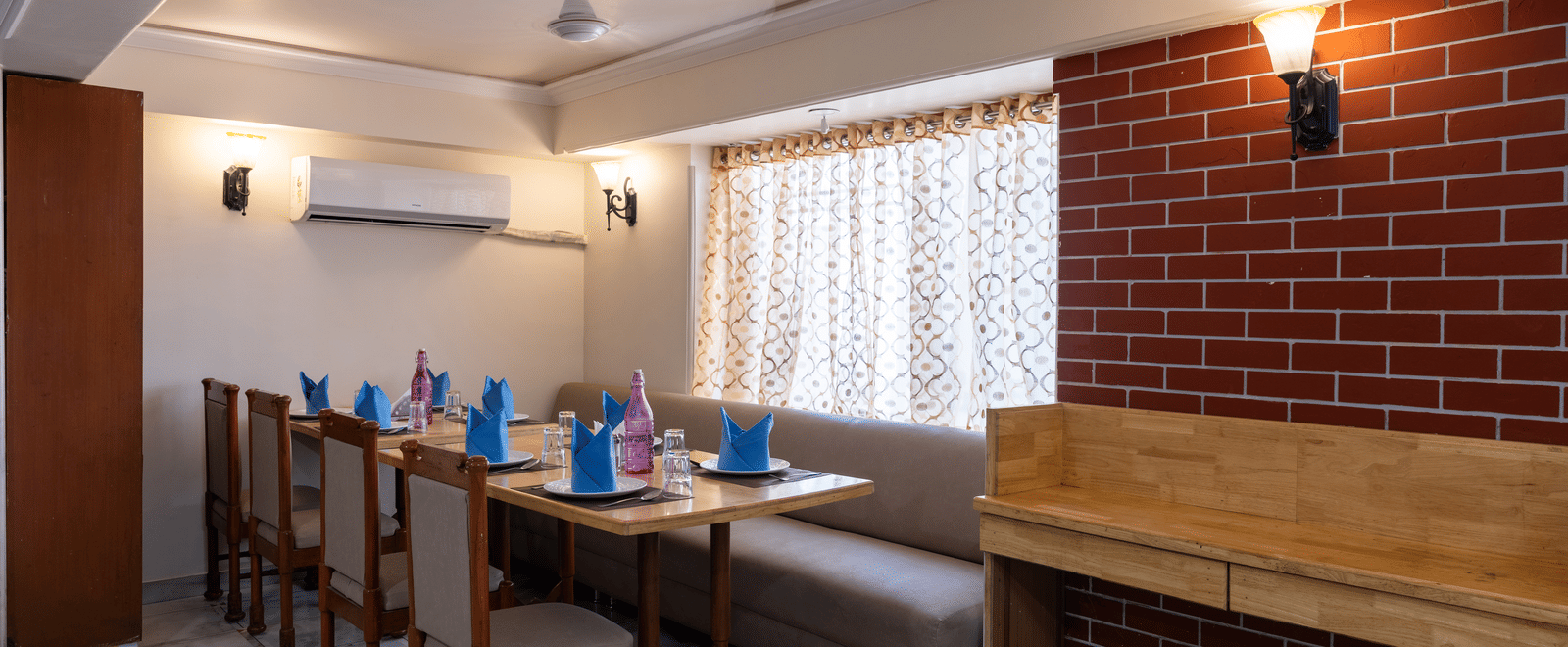 Restaurant seating area featuring wooden chairs, a long booth, exposed brickwork, and an air conditioning unit at Pravasa Stay, Vadodara.