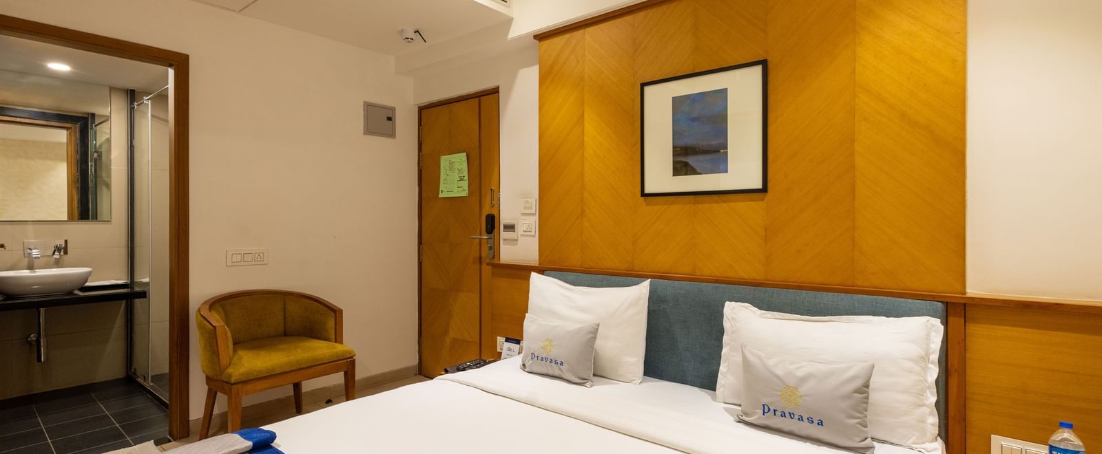 Queen size bed, seating chair, and a door opening to the bathroom seen in a Premium Room of Pravasa Stay Vijay Nagar, Indore.