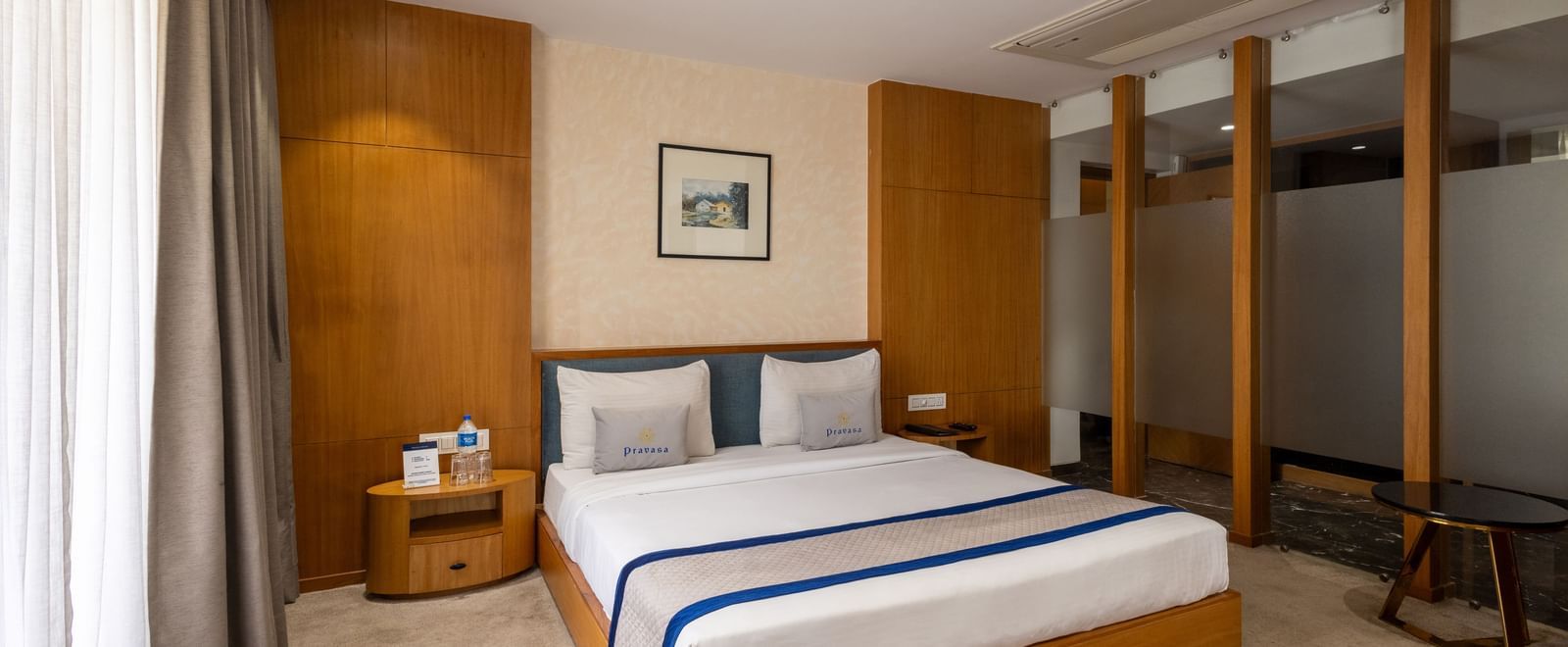 Wide view of the bedroom of Suite at Pravasa Stay Vijay Nagar, Indore, showcasing its queen size bed, bedside tables, and tinted glass partitions.