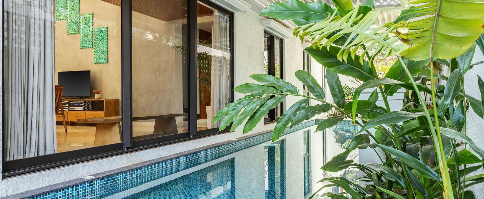 A tranquil swimming pool surrounded by tropical plants, with a glimpse of modern interiors through the glass windows.