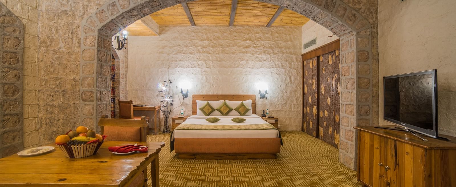 A spacious hotel bedroom with stone walls, wooden furniture, and a welcoming bed under a vaulted ceiling at  Sitara Luxury Hotel in Hyderabad near Ramoji Film City.