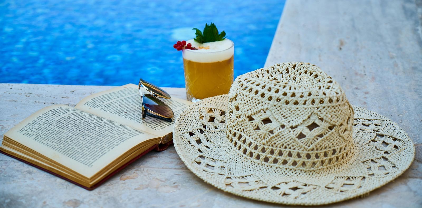A swimming pool with a refreshing juice and a sun hat on the edge.