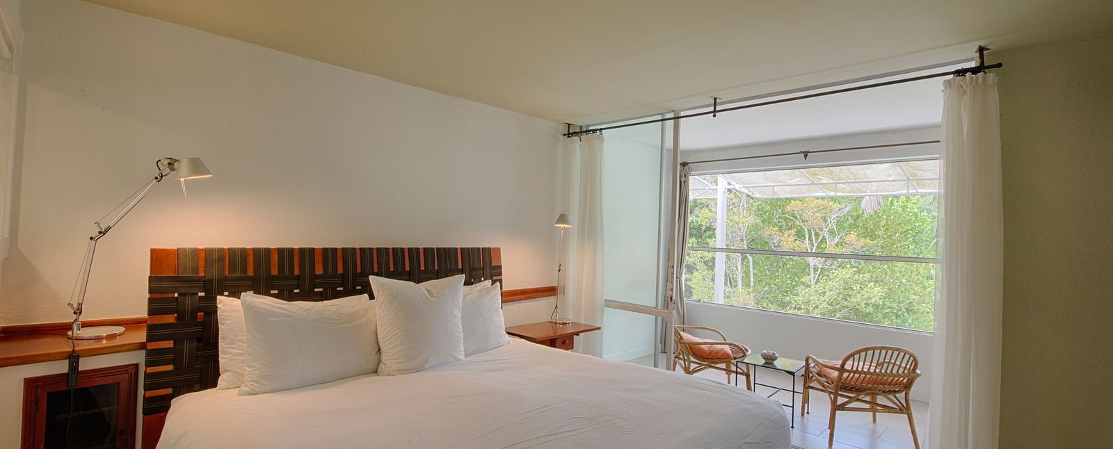 Bed with white linens at Casa Morada, facing a window with a garden view and two chairs.