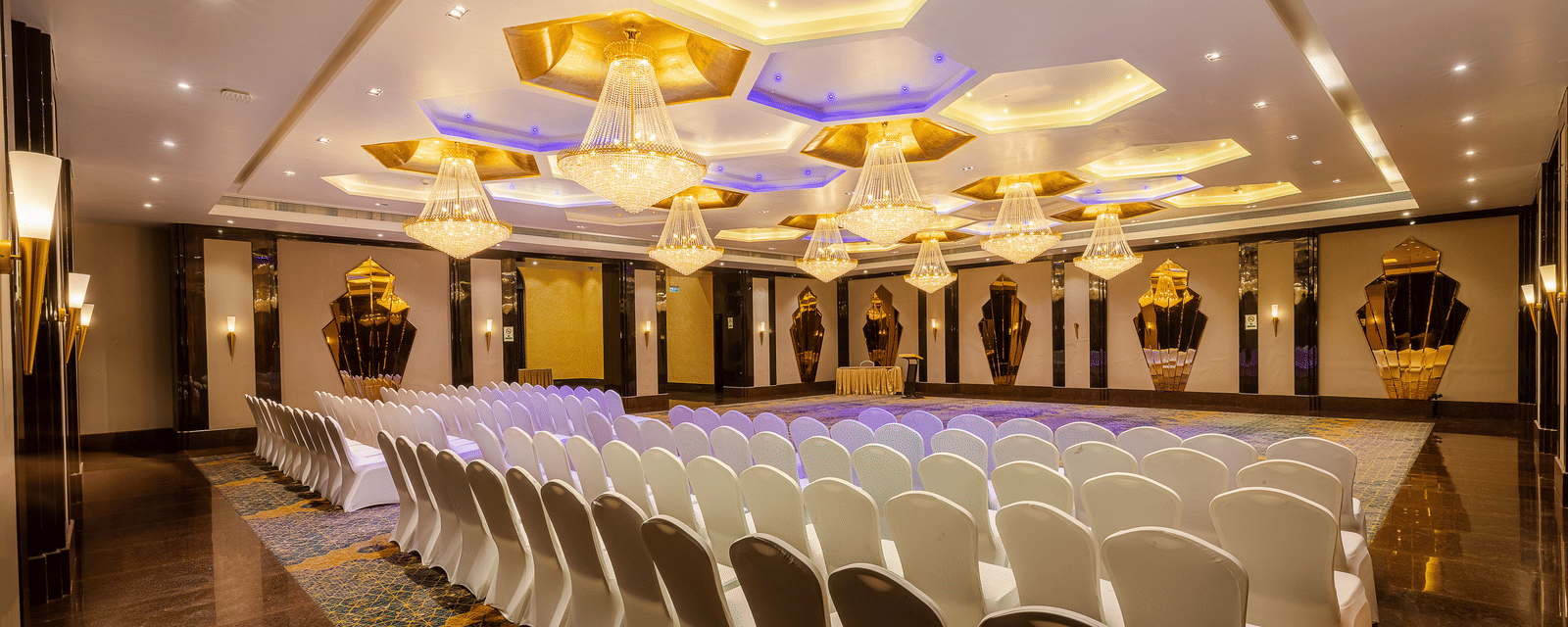 Banquet hall set with rows of white chairs facing a stage area, brightly lit by impressive hexagonal ceiling fixtures at Hotel Hukam's Lalit Mahal.