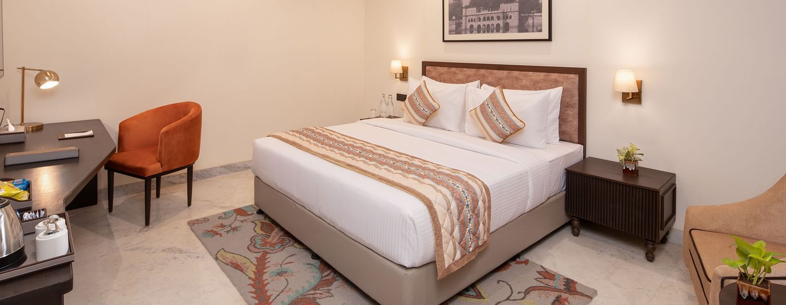 An image of a bedroom with bedside table, photo frame hanged on the wall, work desk with sofa chair, and a flat TV screen - Brij Eternity, Vrindavan