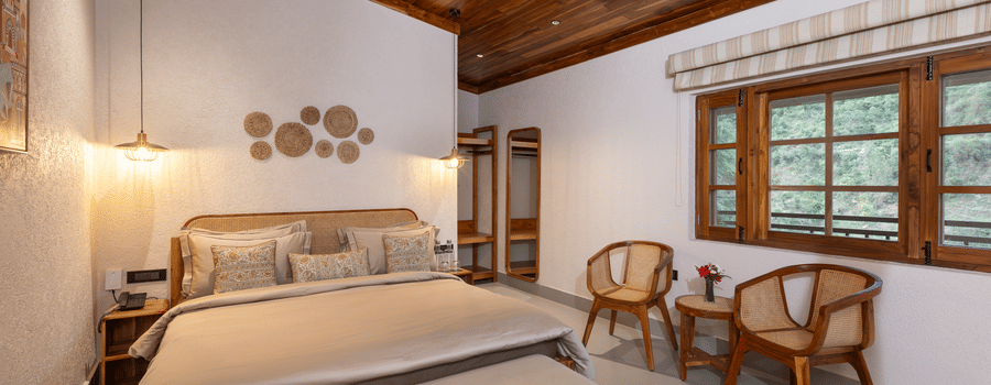 A spacious bedroom with a large bed, two wicker armchairs, a wooden ceiling, and a wide window with blinds | Samsara Hill Resort, Mussoorie