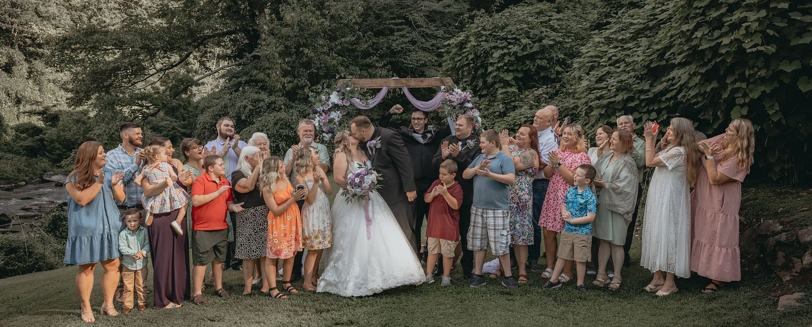 Just married couple kissing each other in front of the wooden frame decorated with flowers and cloth pieces at the Historic Tapoco Lodge. Elders, friends, families, relatives, and kids have also joined their celebration.