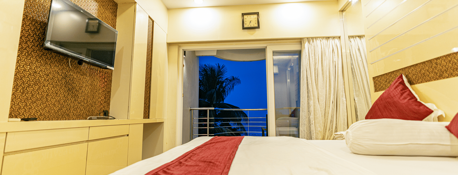 A bedroom with a king size bed with bedding, a television, and a window leading to a balcony at Hotel Seagull Digha.