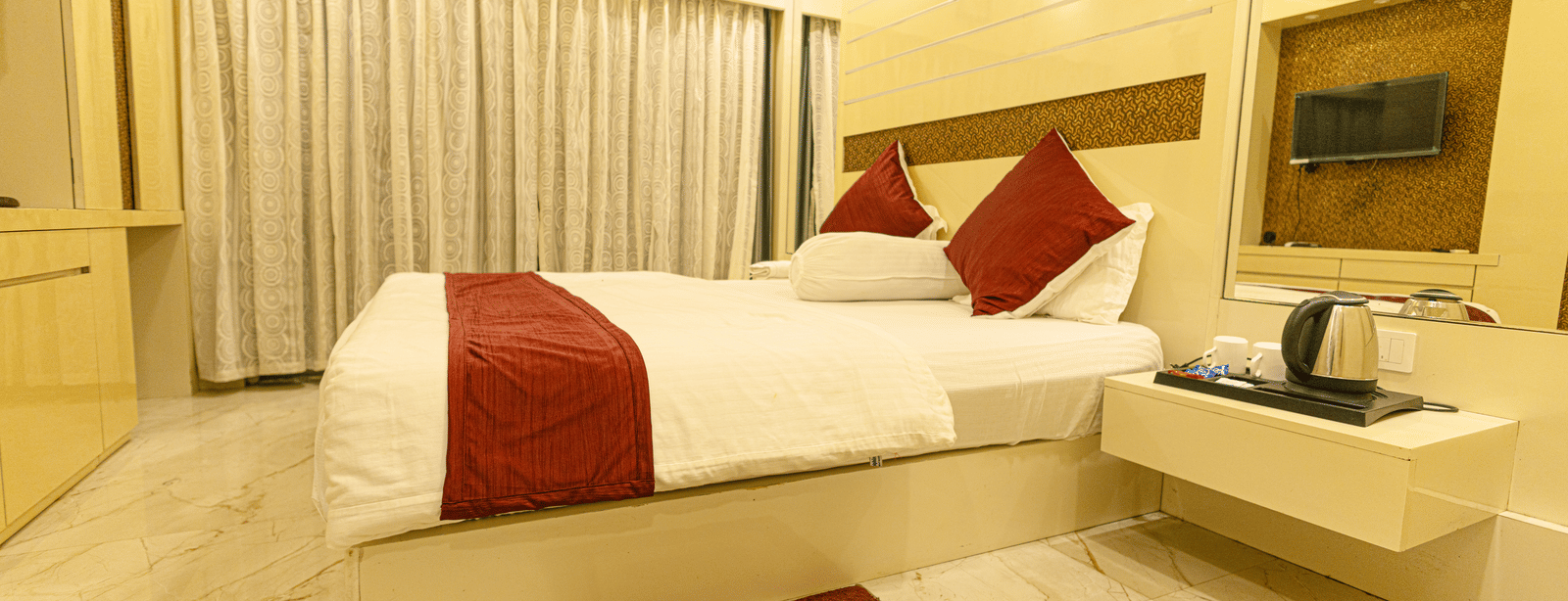 A bedroom with a king size bed, a bedside table with an electric kettle, a television, and curtains at Hotel Seagull Digha.