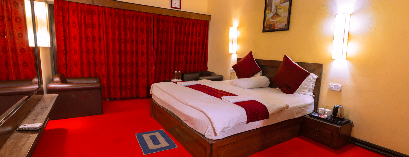 A bedroom with a king size bed, 2 arm chairs, a large mirror, a carpet, a bedside table, wall lamps, and wall art at Hotel Seagull Digha.