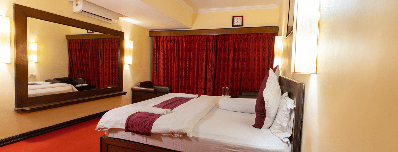 A bedroom with a king size bed, an armchair, a mirror, an A/C, and red carpeting at Hotel Seagull Digha.
