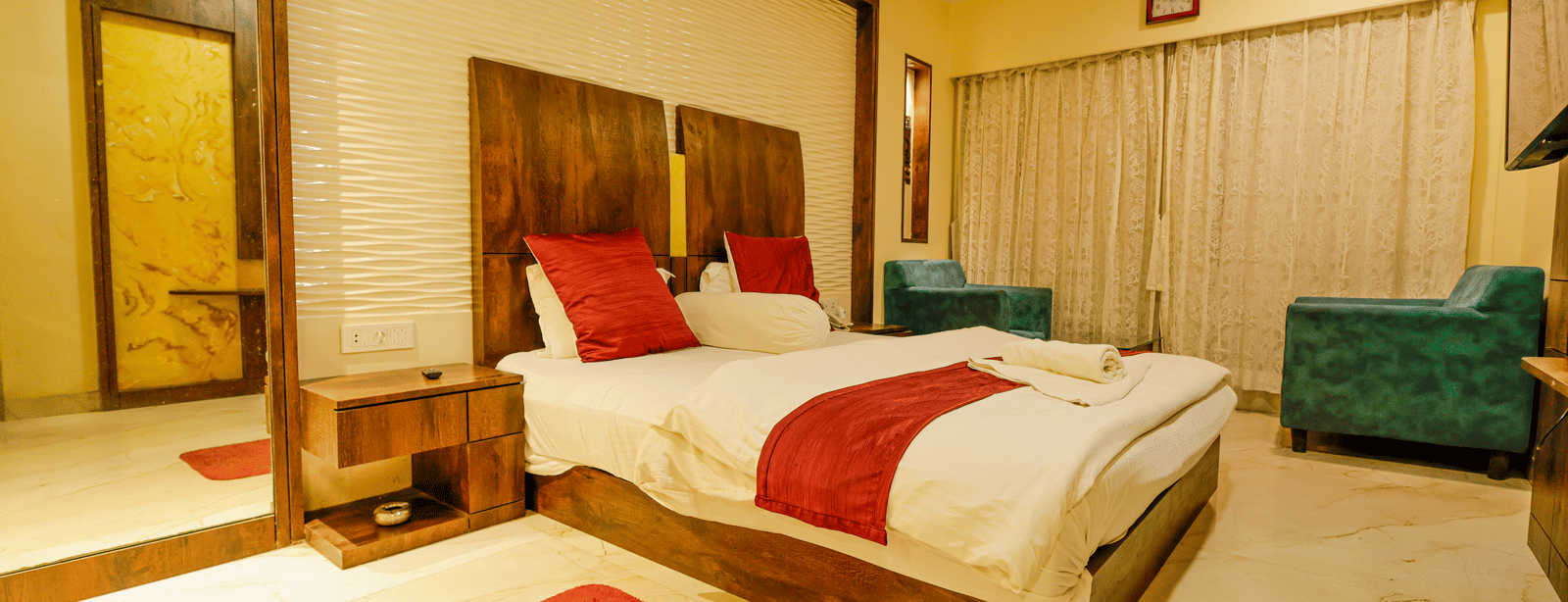 A bedroom with a king size bed, red cushions, a mirror, a bedside table and 2 armchairs at Hotel Seagull Digha.