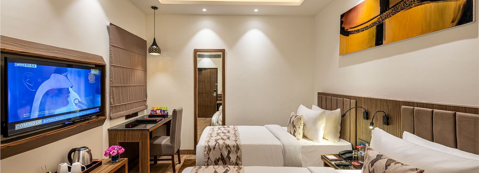 A hotel room with two twin beds, a flat-screen TV, and a work desk. It features a unique tray ceiling with lighting, a ceiling fan, and colourful abstract artwork - Lords Eco Inn Dehradun. 