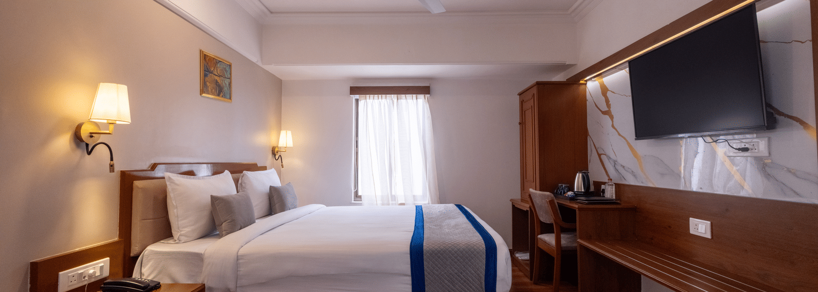 Close-up of the bed and a long wooden desk/console area with a television mounted above the mirror at Pravasa Stay, Vadodara.