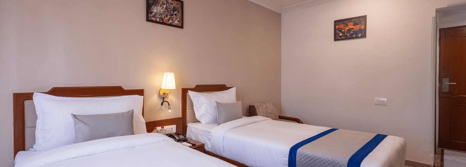 A bright hotel room with two single beds, each made up with white linen and a striking blue and grey throw at Pravasa Stay, Vadodara.