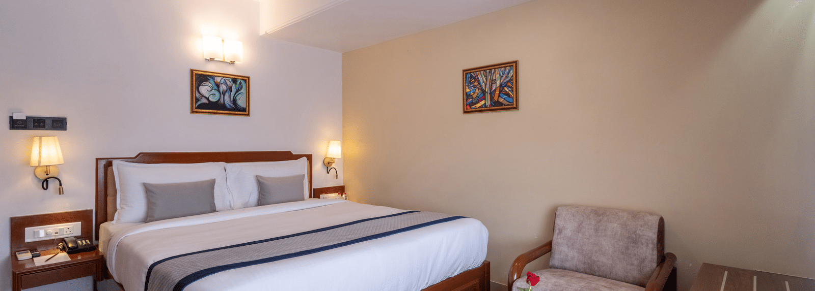 Close-up of a well-made hotel bed and a comfortable armchair with a side table, under ambient wall lights at Pravasa Stay, Vadodara.