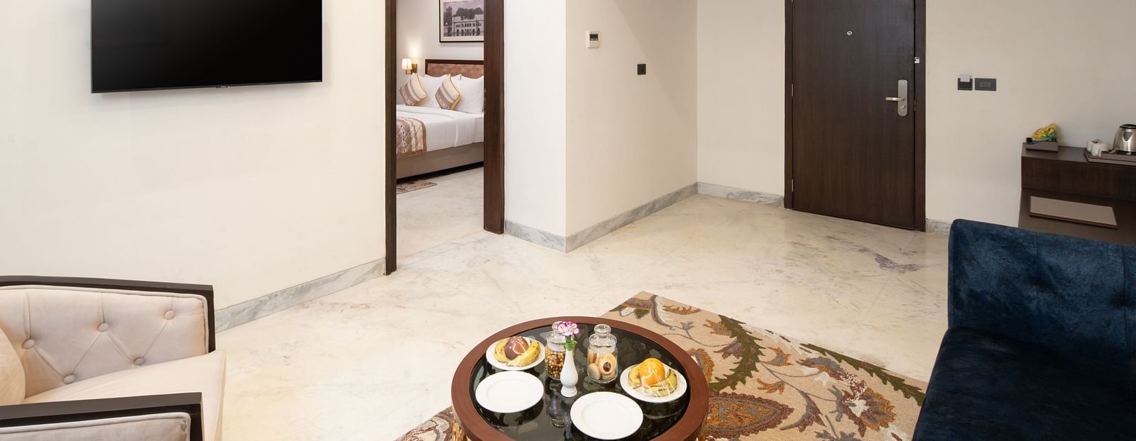 An image of a room with sofa set, centre table, flat TV screen, and a door leading to the next room - Brij Eternity, Vrindavan