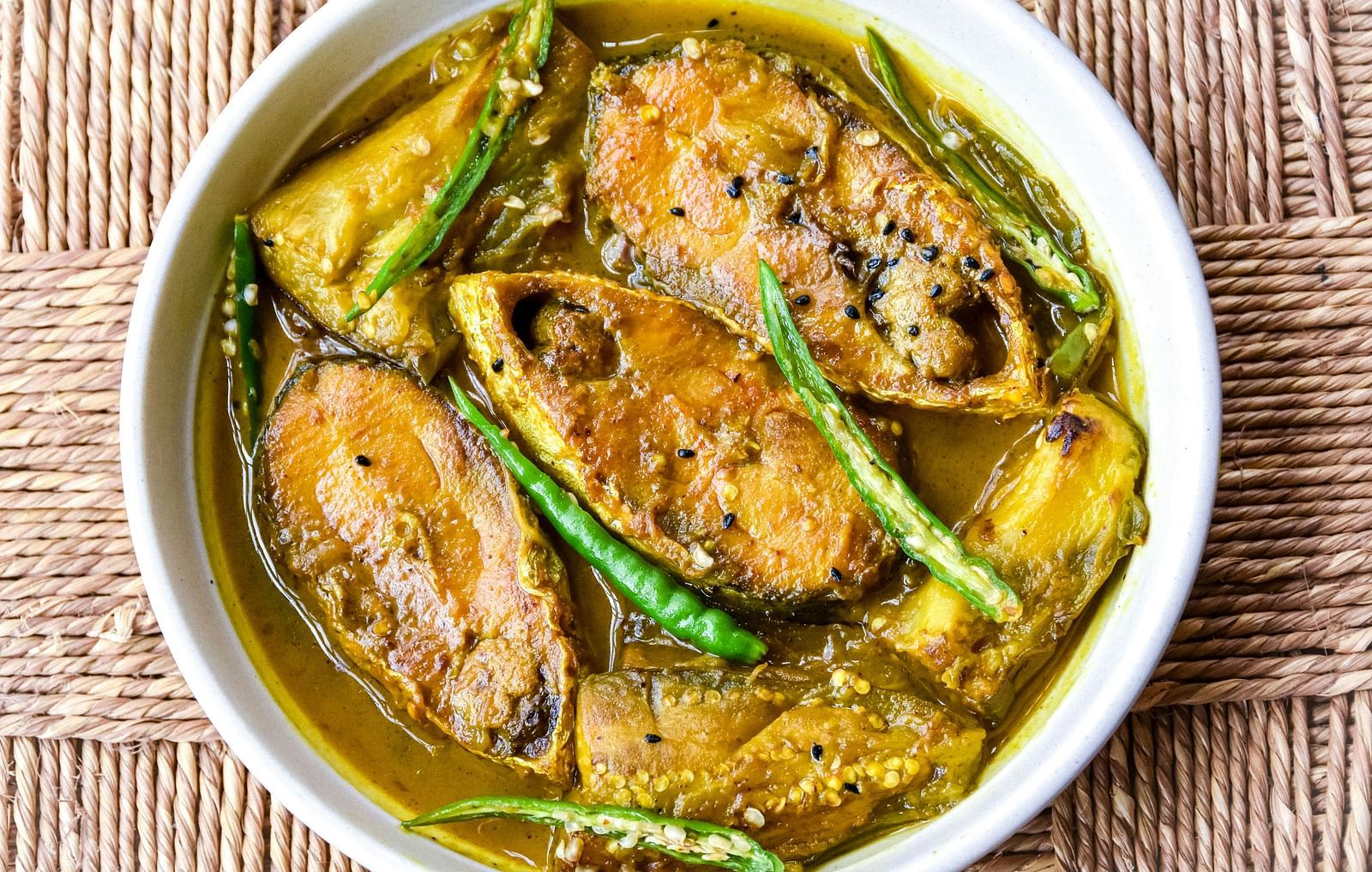 A bowl of fish curry with green chillies on a textured surface.