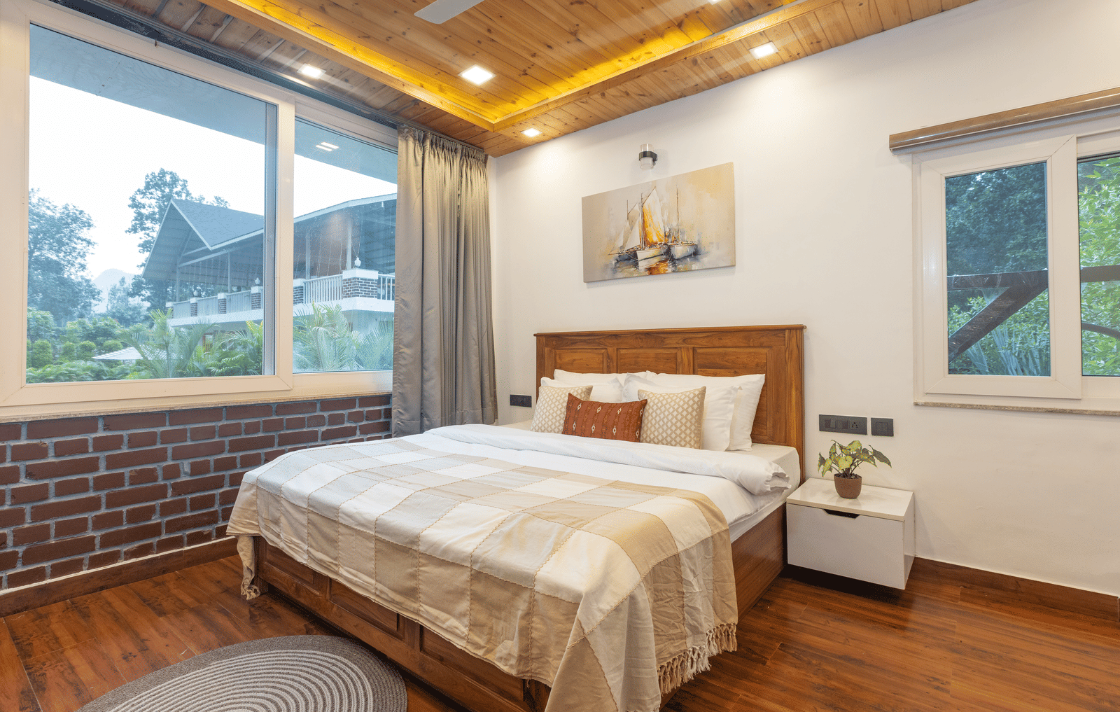 A modern bedroom with a brick accent wall, a large bed, and a window with a scenic view at Megha Resort & Villas, Dehradun.