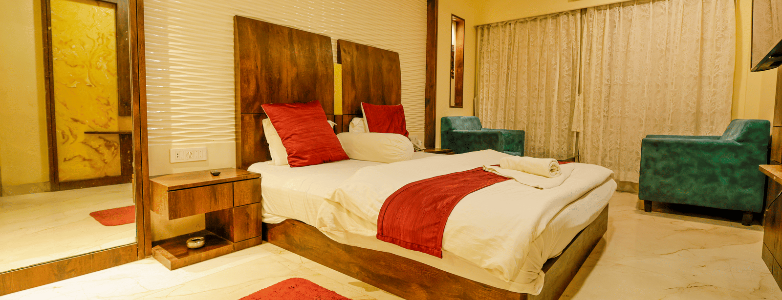 A bedroom with a king size bed, red cushions, a mirror, a bedside table and 2 armchairs at Hotel Seagull Digha.