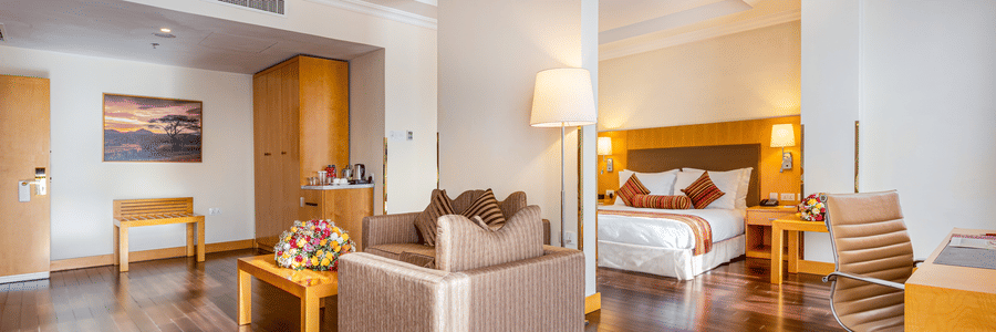 A suite with a bed, seating area, desk, chairs, tables, lamps, wooden floor, wall art, wardrobe, and a tea and coffee setup at Ramada by Wyndham Addis, Addis Ababa.